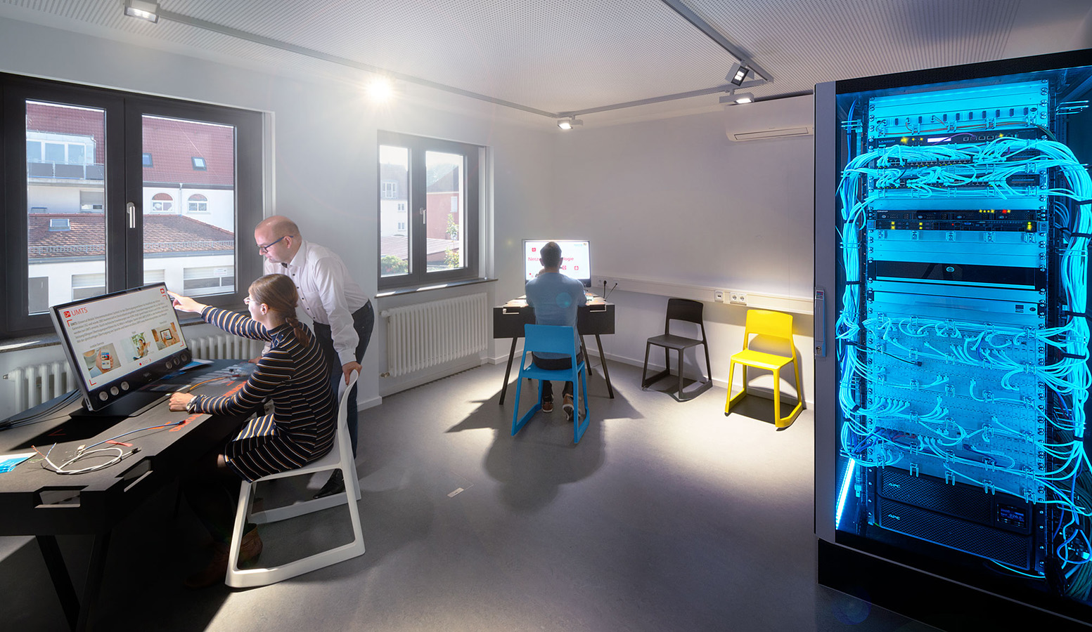 Am linken Bildrand ist ein Computerarbeitsplatz mit zwei Bildschirmen abgebildet an dem zwei Personen arbeiten. In der Mitte steht ein weiterer Computerarbeitsplatz an dem eine Person arbeitet. Rechts im Bild steht ein blau beleuchteter Serverschrank.